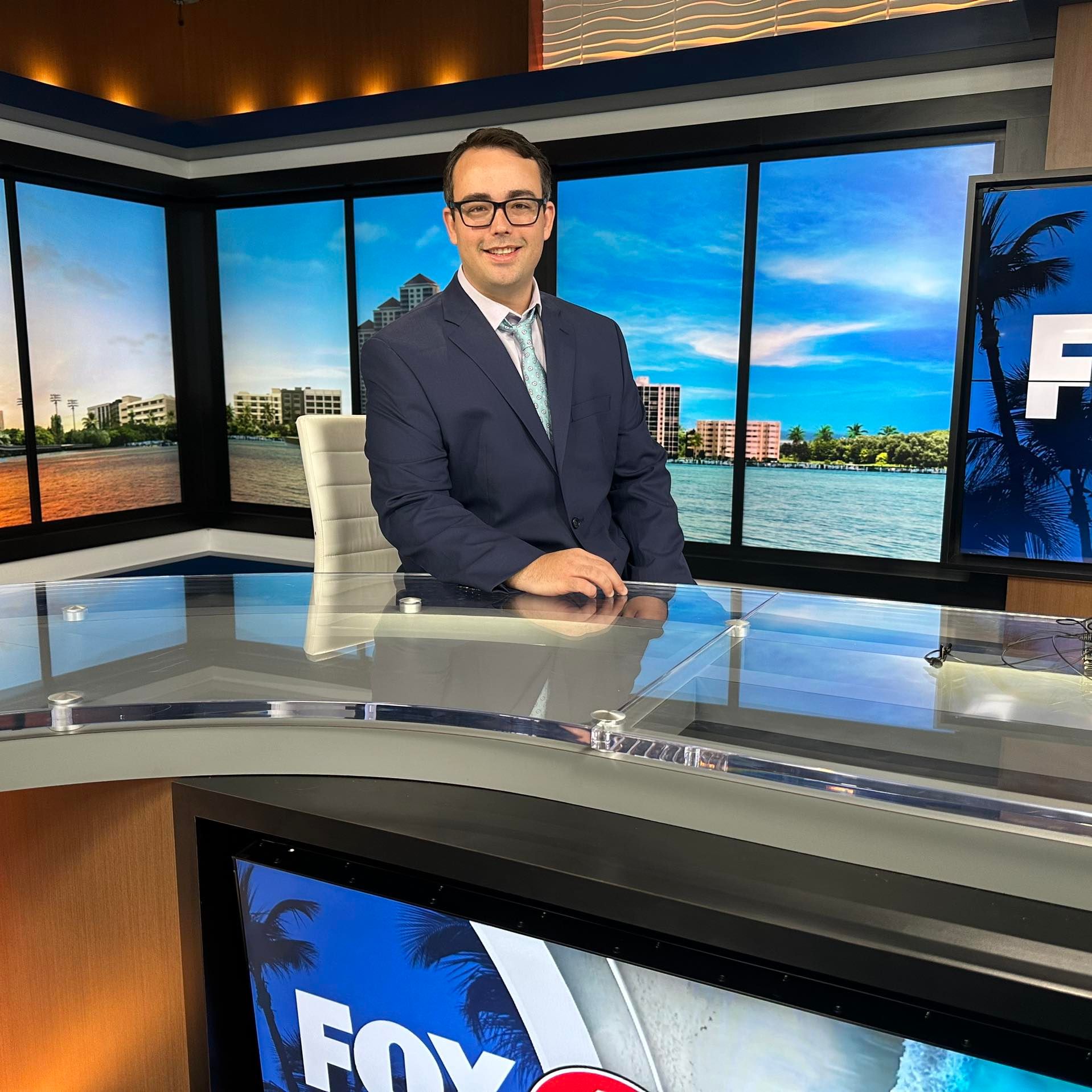 Professional news anchor in a television studio with a scenic waterfront background and the Fox logo on the screen.