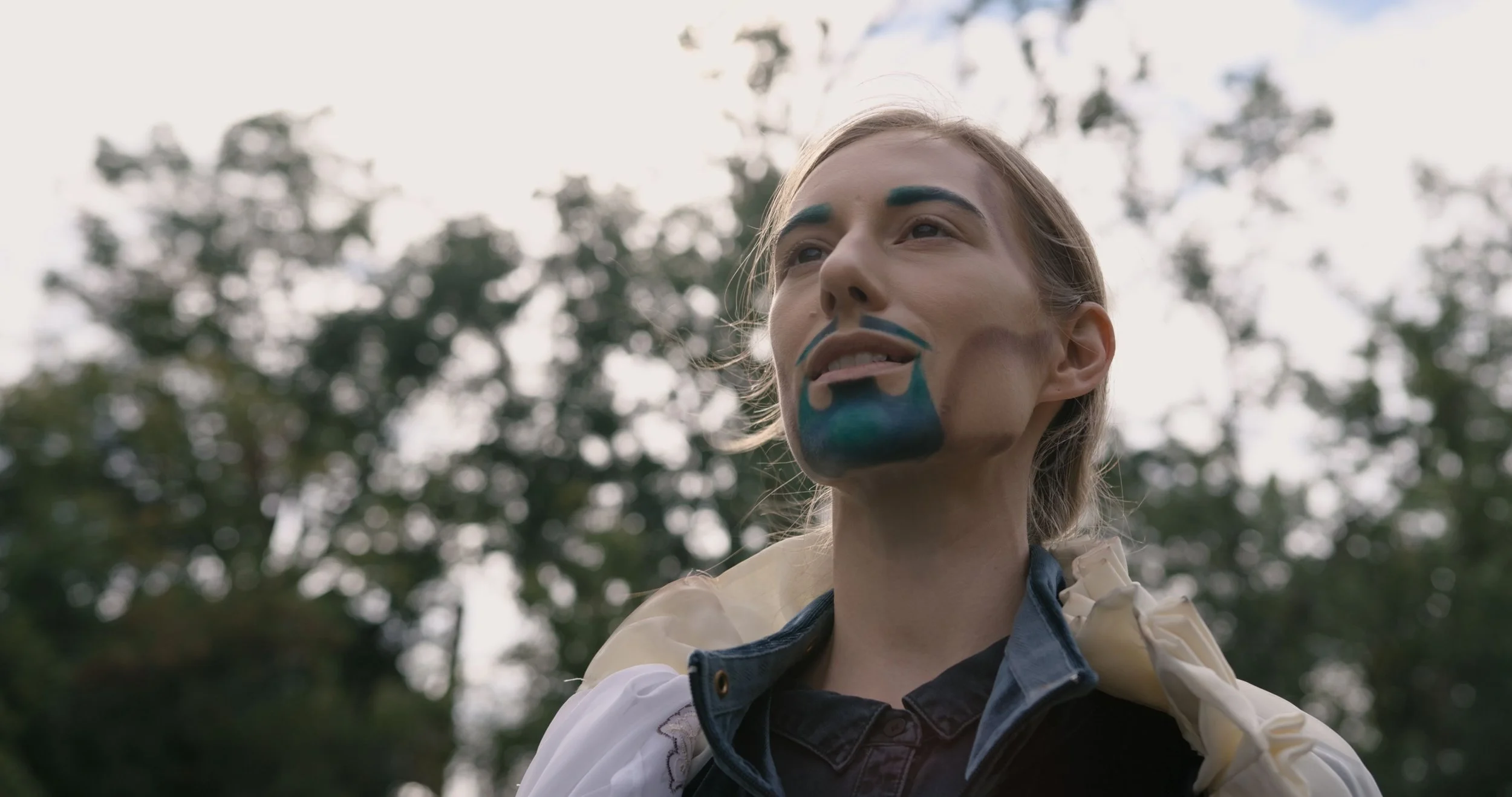 Rachel Miller as Poseidon, with painted blue beard and eyebrows, stands in front of forest background.