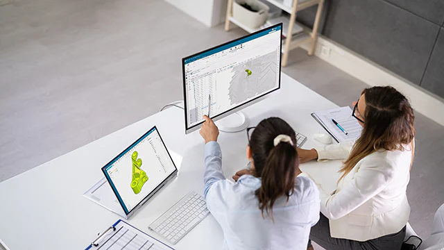 Two female coworkers collaborating at a desk with a computer monitor and tablet displaying Siemens NX CAD software.