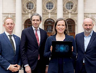 Three men and a woman in business attire stand in a line. The woman holds a tablet that shows the number 550 in blue writing.