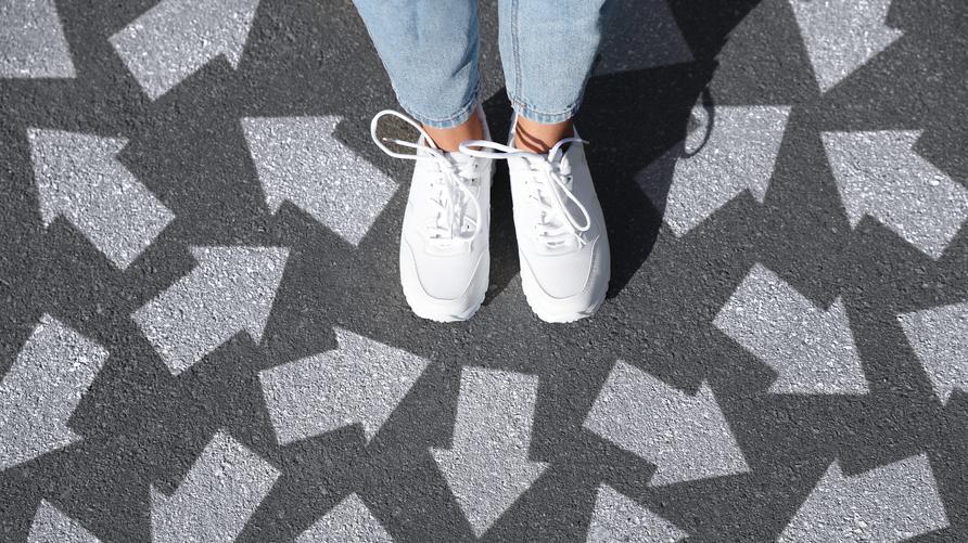 U18-Wahl: BU: Bei der Wahl ist jede Stimme bedeutend, sie bestimmt, wo lang es geht. ++++++++++<br /> Woman standing on asphalt road with white arrows pointing in different directions, top view. Concept of choice and making decisions