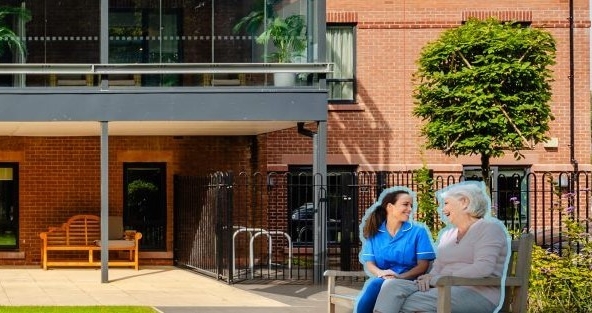 Elderly woman and young woman sitting on a bench in front of a care home smiling at eachother