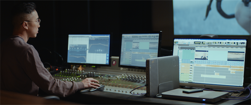 Person working at a sound mixing console with multiple monitors displaying audio waveforms and video editing software, featuring an iodyne Pro Mini SSD for efficient data storage and workflow management.
