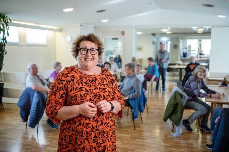 Olaug i front og eldre mennesker bak. Hun er på eldretreff og smiler