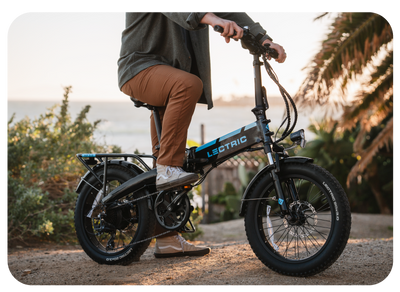 A person wearing a gray jacket and brown pants is riding a black Lectric XP 3.0 e-bike on a dirt path with lush greenery and a beach in the background, framed by the setting sun.