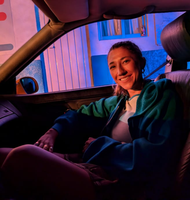 A person sits smiling in a car, in dim lighting, and angled towards the camera.