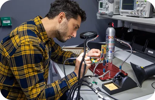 A male Google engineer uses a soldering iron and magnifying glass at his desk for detailed circuitry work.