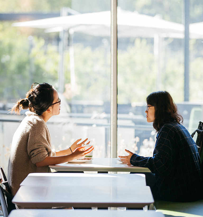 Two women having a conversation a table near a large window.