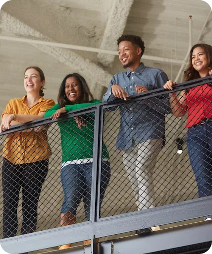 Four Google employees smile while looking down at the lower level of a Google office from a railing above.