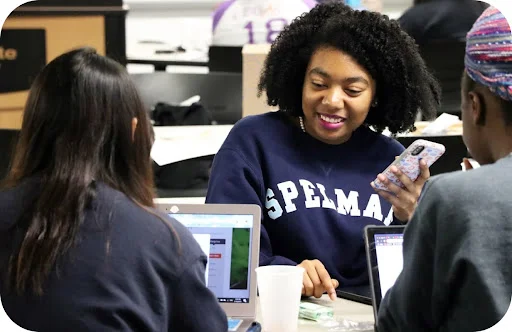 A woman in a Spelman College sweatshirt holds her phone and smiles at two other women around a desk.