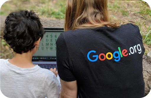 A Google.org fellow sits with a child, both focused on a laptop displaying the alphabet and numbers 1 through 10.