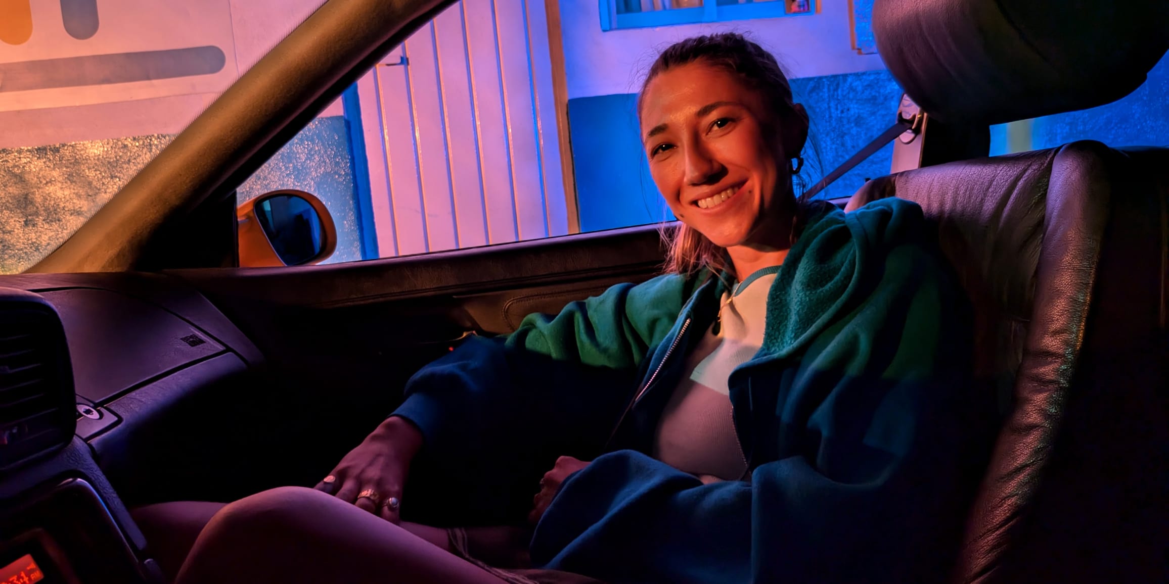 A person sits smiling in a car, in dim lighting, and angled towards the camera.