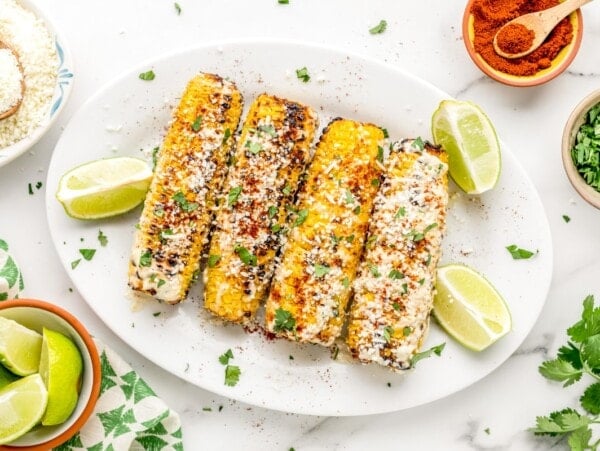 Four ears of Mexican street corn (aka Elote) slathered in cheese and seasonigs, served on a white platter with fresh lime wedges.