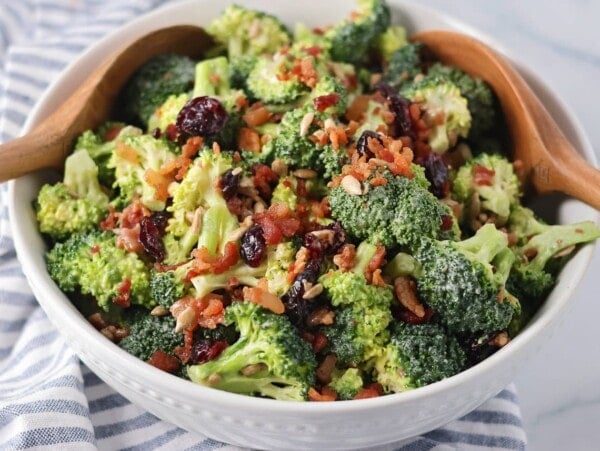 Homemade broccoli salad recipe in white bowl with serving spoons.