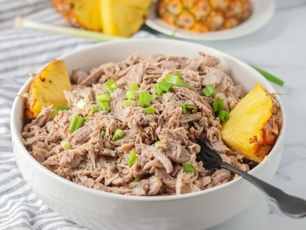 Kalua pork recipe shredded up in white bowl with pineapples on the side.