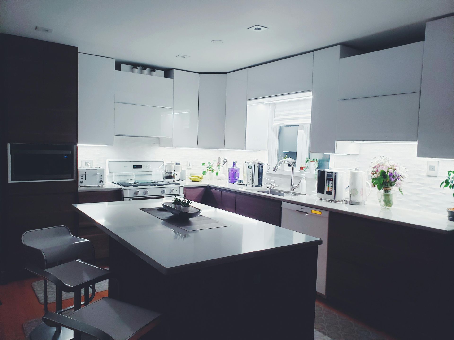 A modern kitchen with dark base cabinets, white upper cabinets, a central island with bar stools, and under-cabinet lighting.