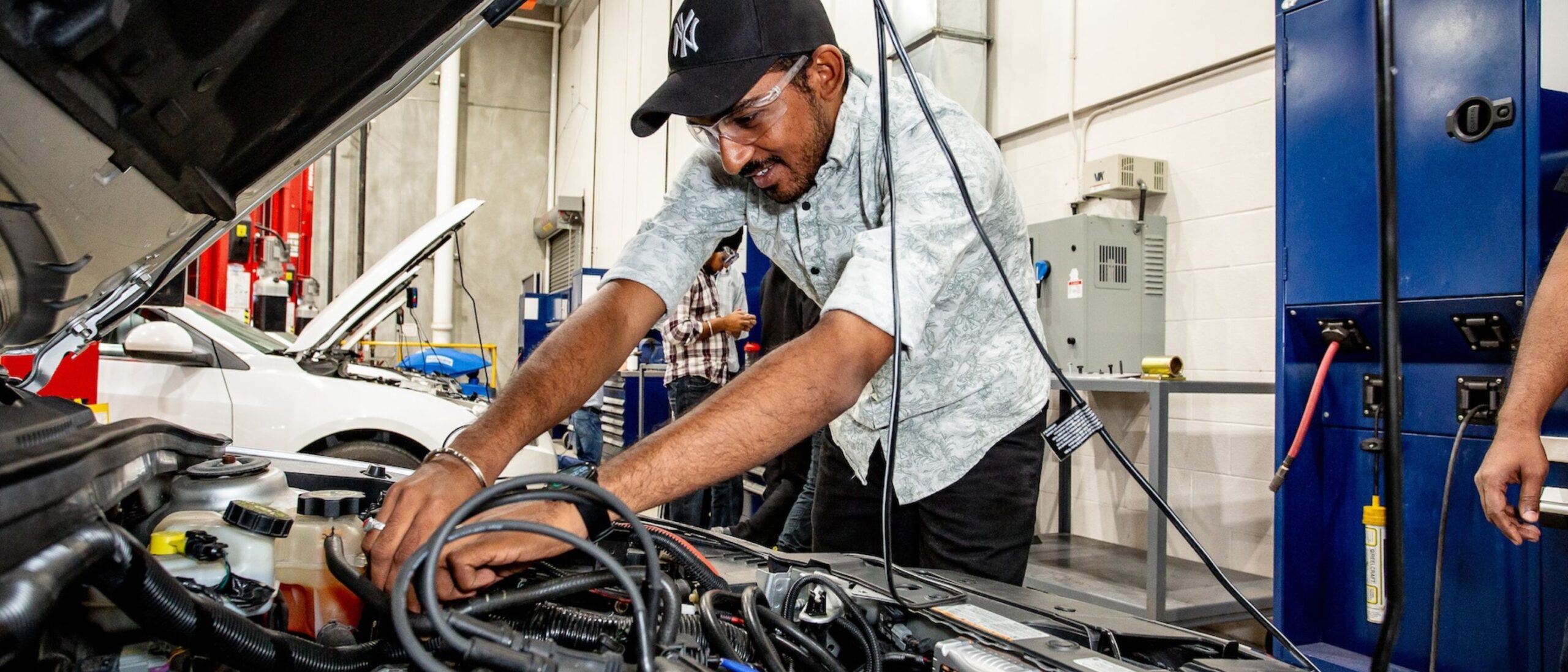 A motive power fundamentals student is working on a car. The student is wearing goggles, short sleeved shirt and a baseball cap.
