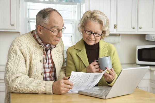 Two people sitting at kitchen table and looking at papers in front of laptop.