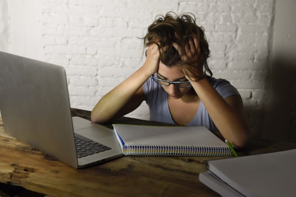 woman with glasses has head down, hands clasped in hair, looking exhausted. Open laptop and blank notebook on desk in front of her woman with glasses has head down, hands clasped in hair, looking exhausted. Open laptop and blank notebook on desk in front of her
