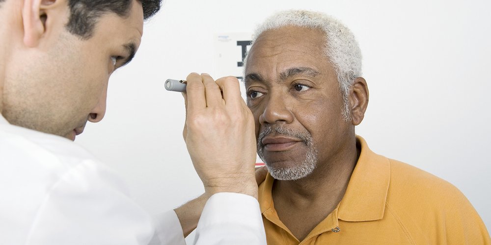 A man having an eye test. He has a focused expression, grey hair and a grey beard and is wearing a pale orange coloured polo shirt. An optician, standing to the left, shines a light from a small torch into his eyes. The optician is looking at the man’s eyes. Behind them is a plain white wall.