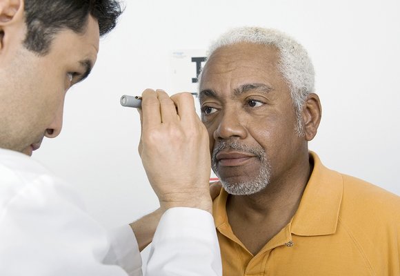 A man having an eye test. He has a focused expression, grey hair and a grey beard and is wearing a pale orange coloured polo shirt. An optician, standing to the left, shines a light from a small torch into his eyes. The optician is looking at the man’s eyes. Behind them is a plain white wall.