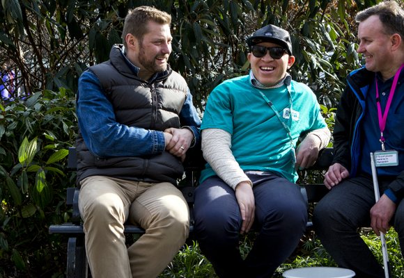 Three men sat on a bench, talking and laughing. One is wearing a blue RNIB shirt and another has a white cane.