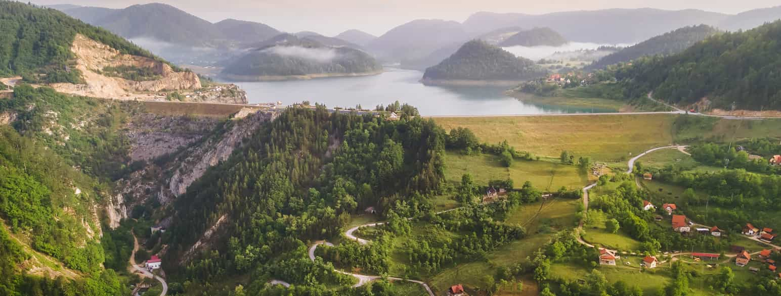 Et grønt fjellandskap med en noen spredte hus og svingete veier opp mot en innsjø. I bakgrunnen skimtes fjelltopper i tåke. Foto