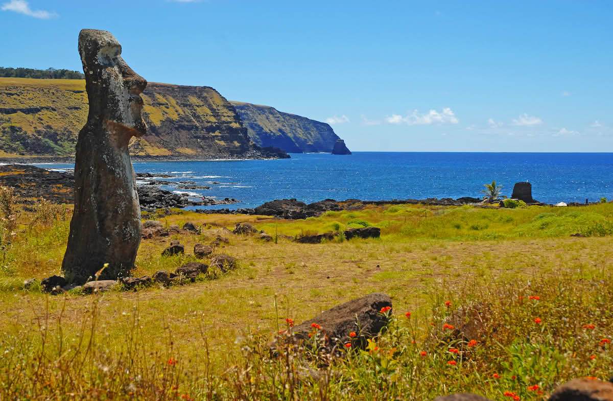 Fotografi av et kystlandskap. I forgrunnen er en stor statue av stein. Landskapet er kupert, og deler av kysten er bratt. Lanskapet er dekket av gress og blomster, men ingen trær.