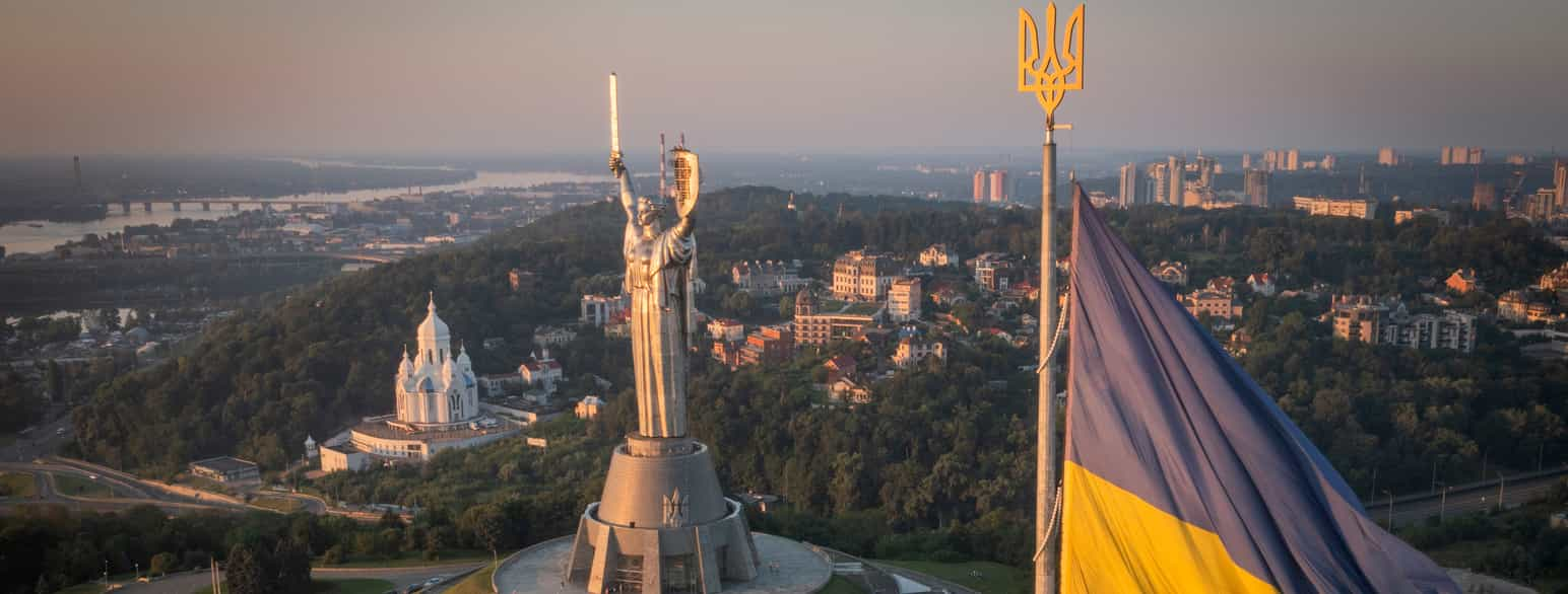 Luftfoto av byen Kiev med en statue av en kvinne som holder opp et sverd og et skjold. I forgrunnen vaier et ukrainsk flagg. Foto