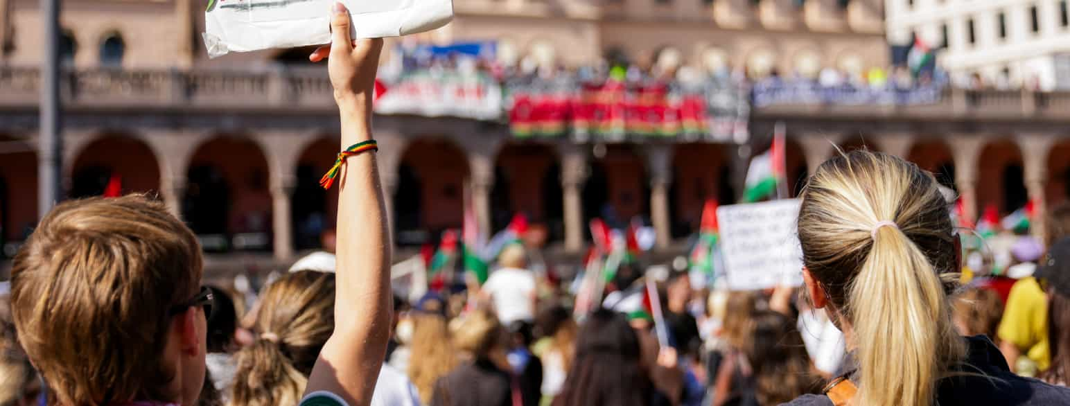 Foto fra et torg med mange mennesker som ser mot en bygning. Bakhodet til to personer er i fokus, den ene holder opp et skilt med den høyre armen. Mange andre mennesker, palestinske flagg, og bygningen med bannere er i ufokus.