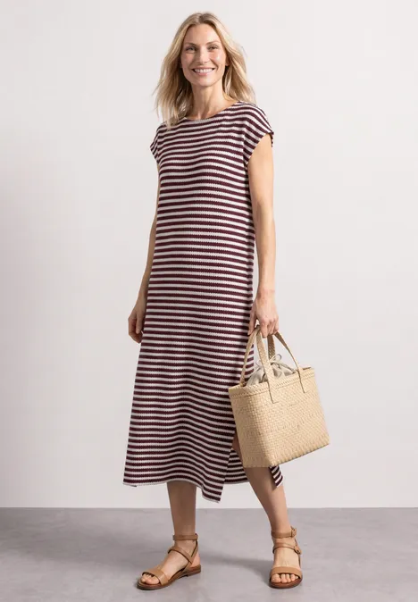 Gebreide jurk rustic rouge Vrouw in gestreepte midi-jurk met sandalen en gevlochten stro-tas tegen een neutrale achtergrond.