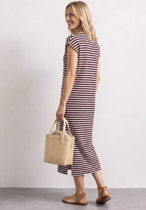 Gebreide jurk rustic rouge Vrouw in gestreepte midi-jurk met rieten tas en platte sandalen, effen achtergrond.