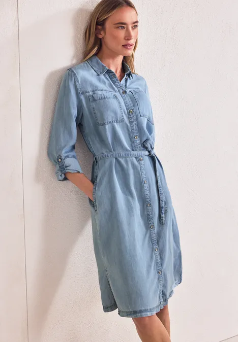 Knielange jurk in een verwassen look Light Blue soft wash Vrouw leunt tegen witte muur in lichtblauwe denimjurk met tailleband en zakken.