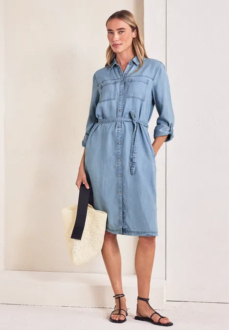 Knielange jurk in een verwassen look Light Blue soft wash Vrouw in lichtblauwe denim jurk met ceintuur, draagt een stro-tas en zwarte sandalen.