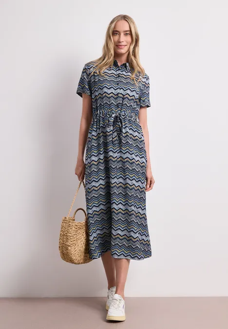 Midi-jurk met zigzagpatroon frozen wave Vrouw in midi-jurk met blauw patroon, strooien tas en witte sneakers voor lichte achtergrond.