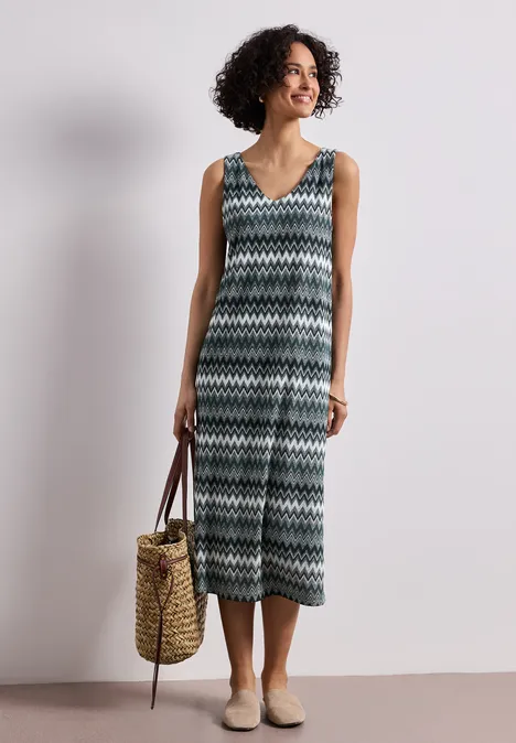 Midi-jurk met V-hals in jacquardstof deep bound green Vrouw draagt mouwloze jumpsuit met V-hals in zwart-wit-groen zigzagpatroon, strozak en beige schoenen.