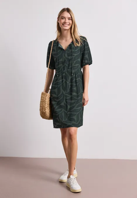 Knielange jurk met gespleten hals en print deep bound green Vrouw in donker groen overhemdjurk met bladprint, witte sneakers en gevlochten strooien tas.