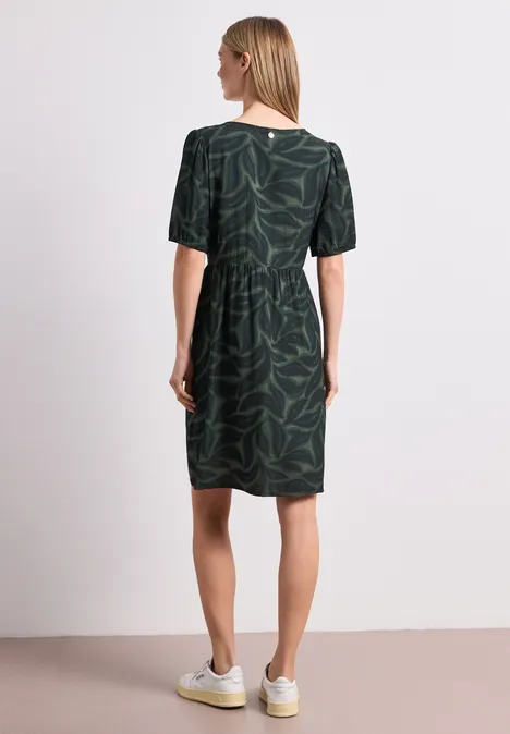 Knielange jurk met gespleten hals en print deep bound green Vrouw van achteren in groene jurk met bladprint en witte sneakers voor witte achtergrond.