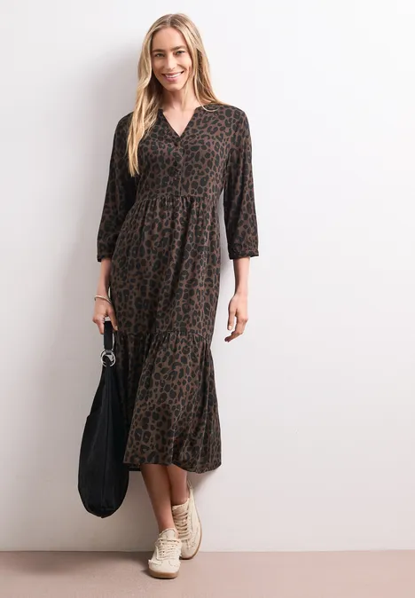 Midi-jurk met gespleten hals en print molten earth Vrouw in midi-jurk met bruin-zwarte print, driekwartmouwen, lichte sneakers en zwarte tas.