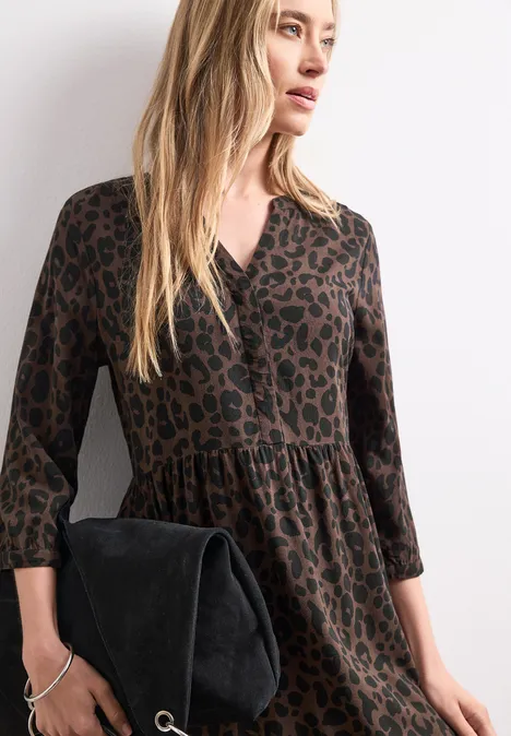 Midi-jurk met gespleten hals en print molten earth Vrouw in bruin luipaardjurk met 3/4-mouwen, zwart suède clutch, lang blond haar.
