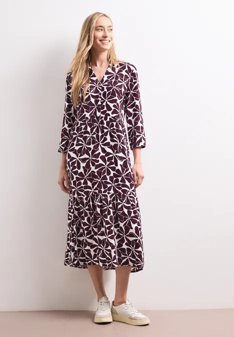 Midi-jurk met gespleten hals en print rustic rouge Blonde vrouw draagt een midi-jurk met wit motief op paars en witte sneakers tegen witte muur.
