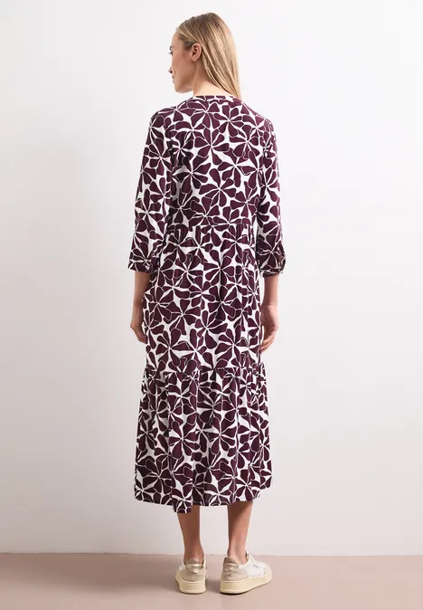 Midi-jurk met gespleten hals en print rustic rouge Rugaanzicht van een vrouw in een paarse midi-jurk met witte bladprint, driekwart mouwen en lichte sneakers.