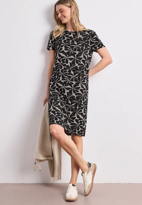 Knielange jurk met pofmouwen Black Vrouw in losvallende, knielange zwarte jurk met witte print, korte mouwen, witte sneakers en lichte jas.
