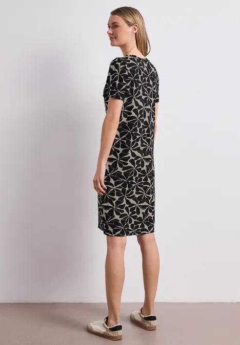 Knielange jurk met pofmouwen Black Vrouw staat in zijaanzicht tegen witte muur, draagt zwarte jurk met wit bladmotief en slip-on sneakers.