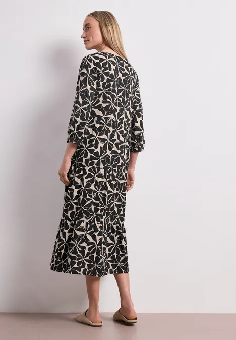 Midi-jurk met gespleten hals in etnische look Black Vrouw staand, rug naar camera, draagt zwarte midijurk met licht bladpatroon en platte sandalen.