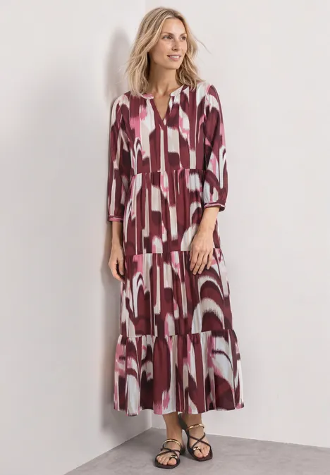 Midi-jurk met gespleten hals in etnische look rustic rouge Een vrouw draagt een lange, losvallende jurk met roze-witte print en driekwartmouwen voor een neutrale achtergrond.