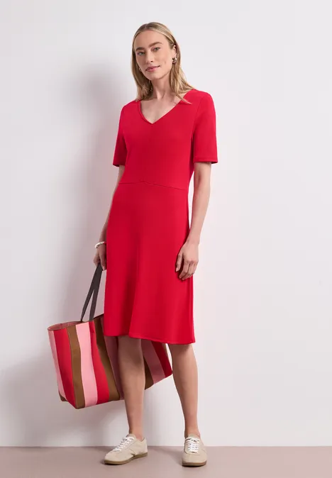 Knielange jersey jurk met korte mouwen salsa red Vrouw in halflange rode jurk met V-hals en korte mouwen, met gestreepte tas en witte sneakers.