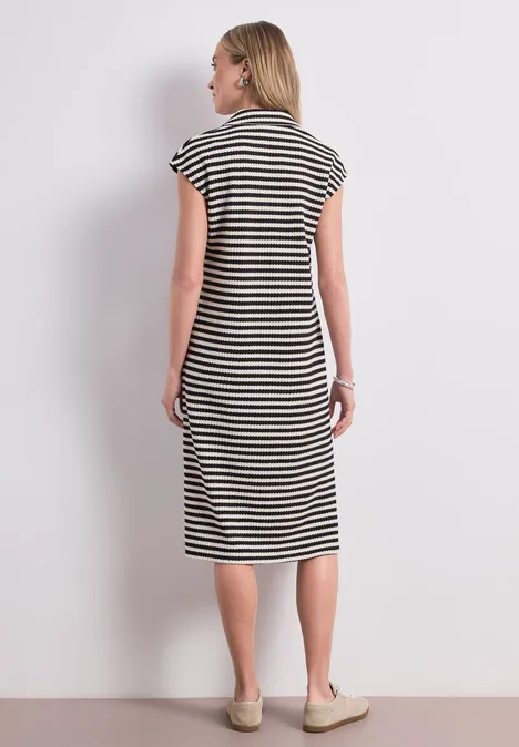 Gebreide midi-jurk met strepen Black Vrouw staat met de rug naar de camera voor een witte muur, draagt een zwart-wit gestreepte midi-jurk en beige schoenen.