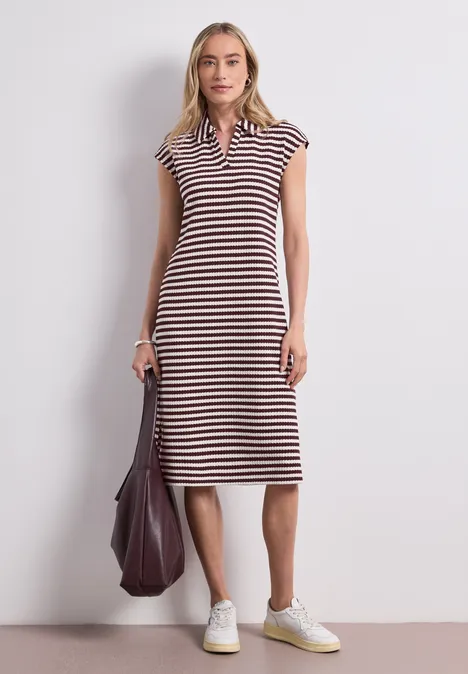 Gebreide midi-jurk met strepen rustic rouge Vrouw in gestreepte midi-jurk met polokraag, witte sneakers en grote tas tegen neutrale achtergrond.