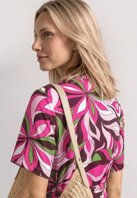 Midi-jurk met V-hals en print magenta dream Blonde vrouw draagt een kleurrijk t-shirt met bloemenpatroon en een strooien tas over haar schouder.